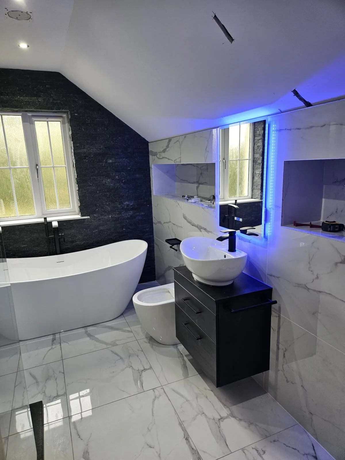 Ensuite fitted by Premier Plumbing — freestanding bath against split-face black stone, marble tiling and a blue-lit LED mirror